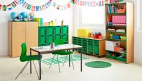 Wooden table, storage cupboards and bookcases in a nursery with alphabet bunting and green plastic chairs