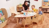 Nursery teacher and children sat on wooden chairs at a semi circular wooden table colouring