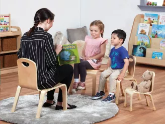 Nursery teacher sat on wooden chair reading a book to boy and girl sat on chairs.