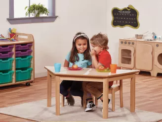 Boy and girl sat on wooden chairs at a table in a nursery with storage behind.