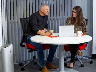 Man and woman sat at round meeting room table with laptop, notebooks and cups of coffee
