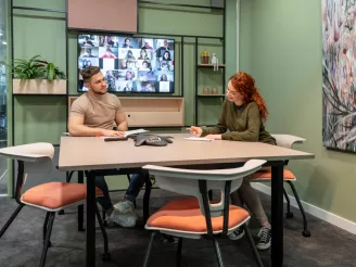Millenial office workers sat at meeting room table with wall mounted screen