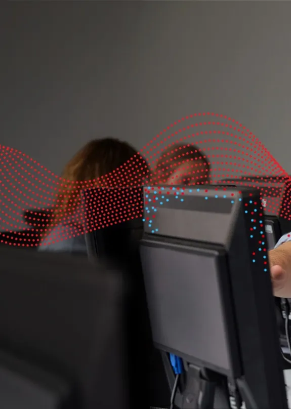 A pupil (girl) sat looking at a desktop monitor in a classroom as her teacher stands beside her as he helps her with her work