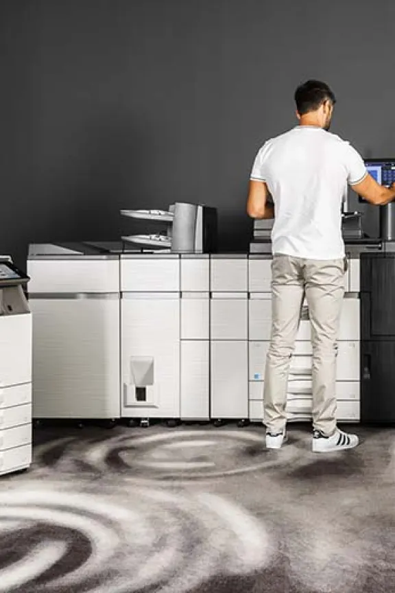 Man wearing white shirt and jeans standing next to a light production printer whilst operating it